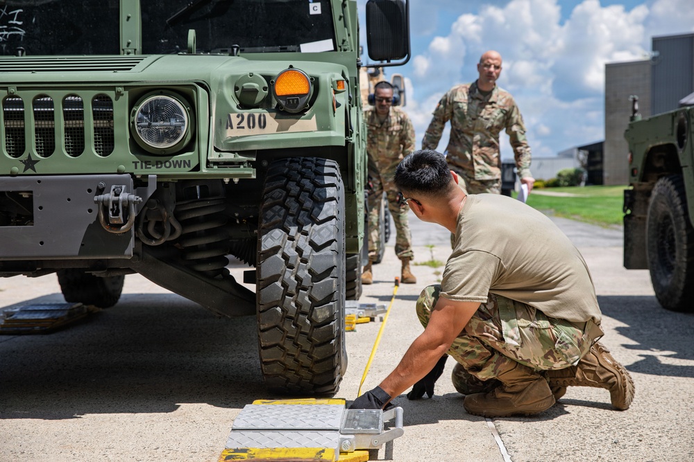 Military personnel in OCP (Operational Camouflage Pattern) uniforms are conducting a vehicle inspection or preparation activity around a military HMMWV (Humvee) marked 'A200'. One soldier is crouched near the front tire, handling yellow measurement tape or tie-down equipment, while two other soldiers approach from behind. The setting appears to be a military installation or airfield tarmac on a clear, sunny day.