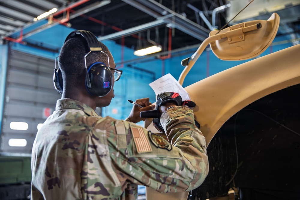 A U.S. military service member in OCP (Operational Camouflage Pattern) uniform is conducting an inspection or maintenance check on a tan/desert-colored military vehicle inside a maintenance bay or hangar. The soldier is holding a document and a writing instrument, appearing to record observations or complete a checklist. The setting is an indoor military maintenance facility with industrial overhead infrastructure visible.