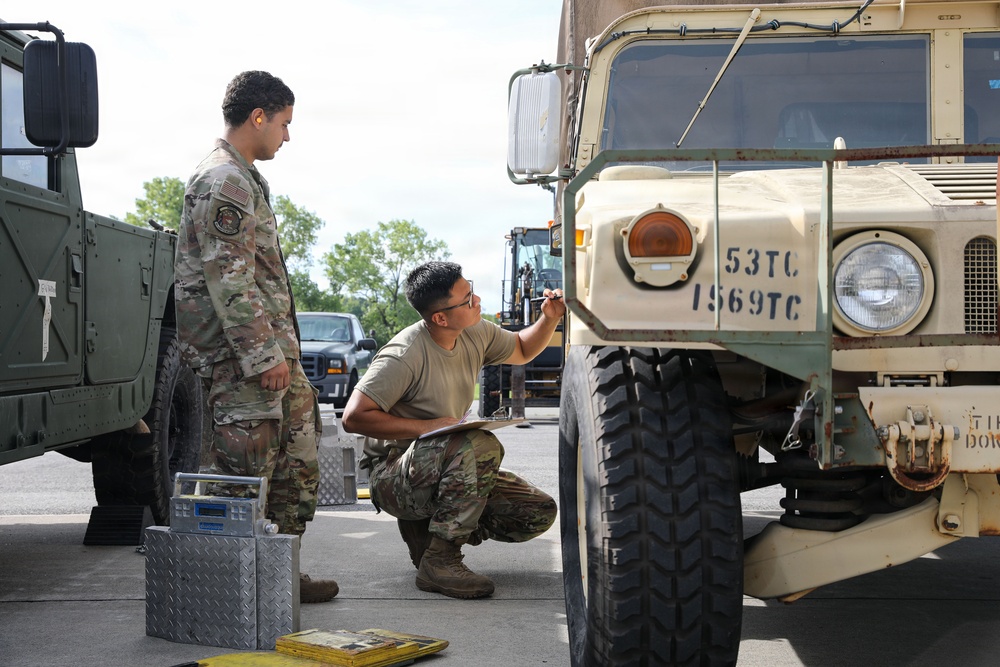 Two military personnel in U.S. Army OCP (Operational Camouflage Pattern) uniforms are conducting an inspection or inventory of military vehicles in an outdoor staging area. One soldier is crouching and writing on a clipboard while marking or noting information on the front of a tan military truck marked '53TC / 1569TC'. The scene appears to be a vehicle maintenance or pre-deployment equipment check.