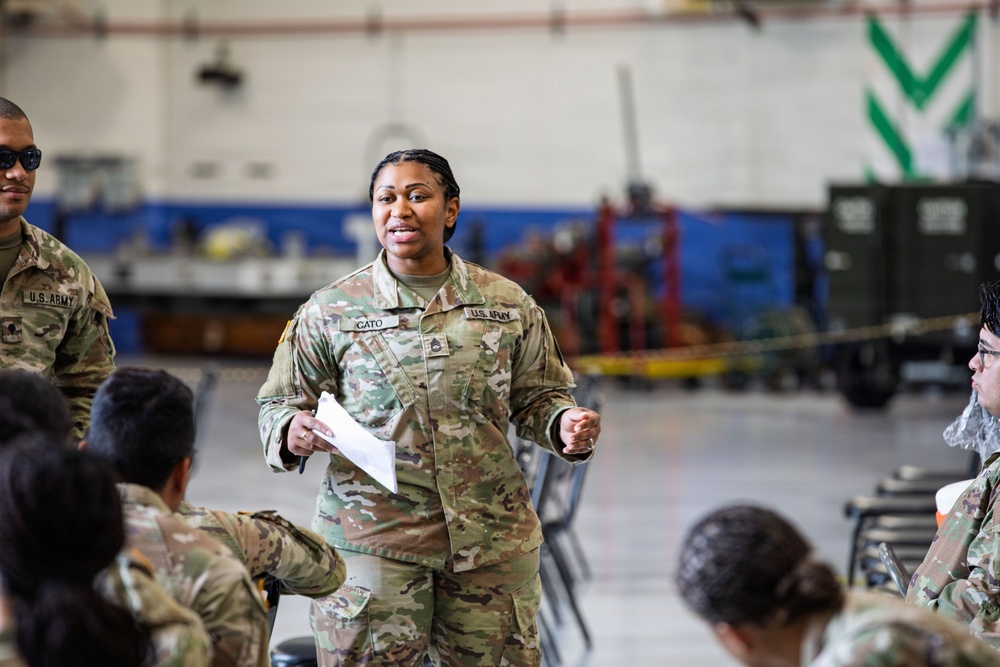 A U.S. Army soldier with the name tape 'CATO' and sergeant rank insignia is addressing a group of fellow soldiers in what appears to be a military maintenance hangar or motor pool facility. She is holding papers and appears to be conducting a briefing or training session. Multiple soldiers in OCP (Operational Camouflage Pattern) uniforms are seated and listening.