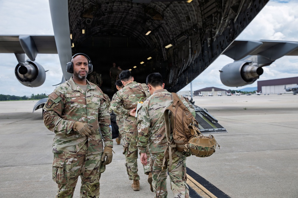 Military personnel in OCP (Operational Camouflage Pattern) uniforms are boarding or loading a large military transport aircraft, likely a C-17 Globemaster III, on a flight line tarmac. One airman in the foreground is wearing aviation headset/hearing protection and tactical gloves, facing the camera. A military hangar and additional aircraft are visible in the background.