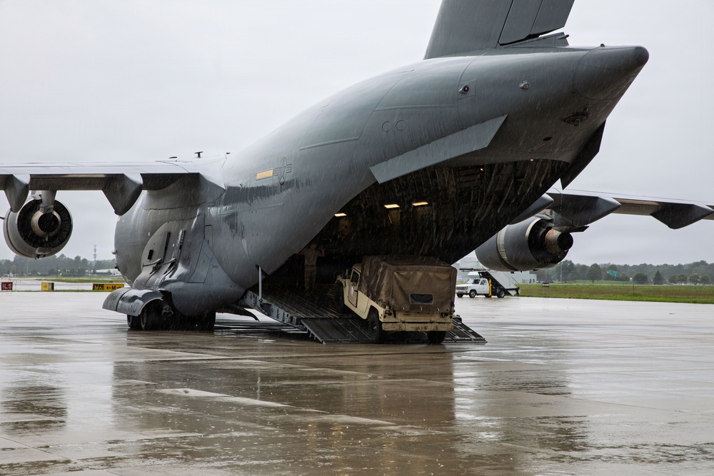 A large U.S. military cargo aircraft, identifiable as a C-17 Globemaster III, has its nose visor raised and front loading ramp deployed on a wet tarmac during rainy conditions. A military Humvee (HMMWV) is in the process of being loaded or unloaded through the aircraft's distinctive visor-nose front cargo door. The scene appears to be at a U.S. Air Force base, with ground support vehicles visible in the background.