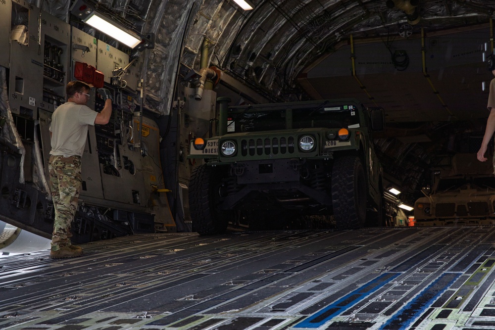 The image shows the interior of a large military cargo aircraft, likely a C-17 Globemaster III, with a military HMMWV (Humvee) being loaded or secured inside. A soldier in MultiCam/OCP camouflage uniform is working on securing equipment along the aircraft's interior wall. Additional military vehicles are visible further back in the cargo hold.