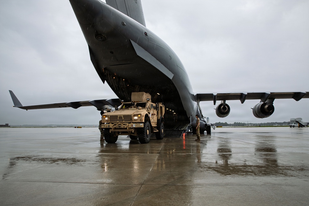 A tan/desert-camouflage MRAP (Mine-Resistant Ambush Protected) vehicle is being offloaded or loaded onto a large military cargo aircraft, likely a C-17 Globemaster III, via its rear ramp. The operation is taking place on a wet tarmac under overcast, rainy conditions. At least two military personnel are visible guiding the vehicle during the operation.