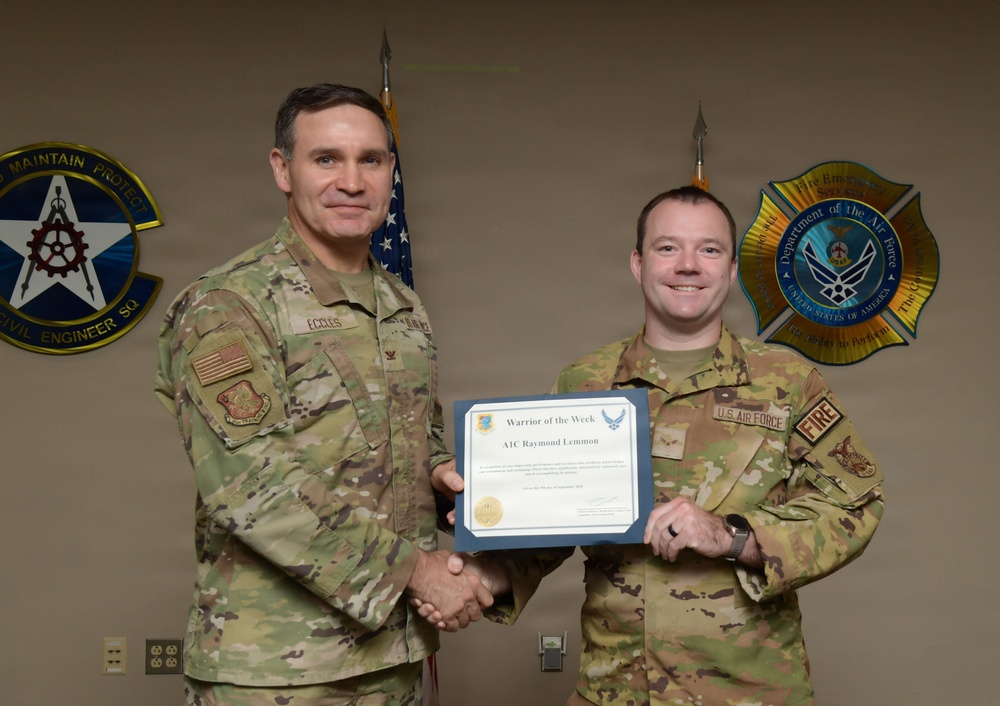 Two U.S. Air Force personnel in OCP (Operational Camouflage Pattern) uniforms are posing together while holding a 'Warrior of the Week' certificate awarded to A1C Raymond Lemmon. The individual on the left, identified by name tape as 'Eccles,' appears to be a senior officer presenting the award. The photo is taken in front of unit emblems including a Civil Engineer Squadron patch and a Department of the Air Force Fire Emergency Services emblem.