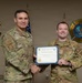 Two U.S. Air Force personnel in OCP (Operational Camouflage Pattern) uniforms are posing together while holding a 'Warrior of the Week' certificate awarded to A1C Raymond Lemmon. The individual on the left, identified by name tape as 'Eccles,' appears to be a senior officer presenting the award. The photo is taken in front of unit emblems including a Civil Engineer Squadron patch and a Department of the Air Force Fire Emergency Services emblem.