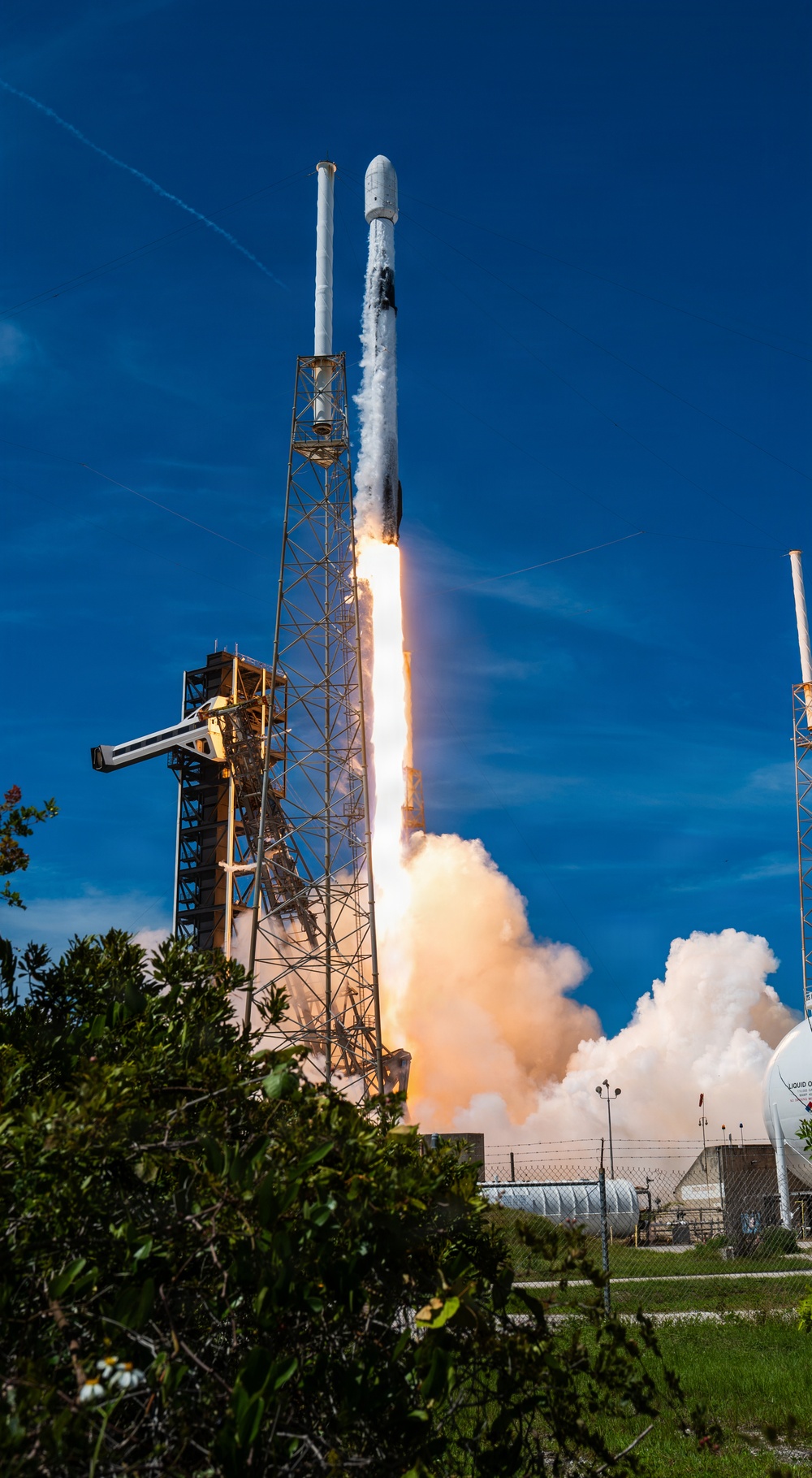 A rocket is captured at the moment of liftoff from a launch pad, with intense flames and billowing white and orange exhaust clouds visible beneath the vehicle. The rocket, identifiable by its white body and payload fairing, is ascending vertically from a steel launch structure. The scene is set against a clear blue sky with green vegetation visible in the foreground.