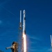 A rocket is captured at the moment of liftoff from a launch pad, with intense flames and billowing white and orange exhaust clouds visible beneath the vehicle. The rocket, identifiable by its white body and payload fairing, is ascending vertically from a steel launch structure. The scene is set against a clear blue sky with green vegetation visible in the foreground.