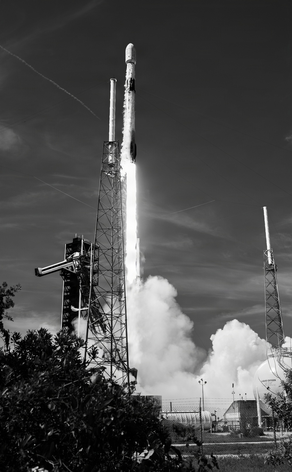 A black-and-white photograph captures a rocket at the moment of liftoff from a launch pad, with massive plumes of exhaust and steam billowing outward. The rocket, consistent in shape with a SpaceX Falcon 9, is ascending vertically with a bright exhaust column beneath it. Ground infrastructure including a launch tower, lightning rod masts, and a visible 'LIQUID OXYGEN' storage tank are present in the foreground and background.