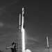 A black-and-white photograph captures a rocket at the moment of liftoff from a launch pad, with massive plumes of exhaust and steam billowing outward. The rocket, consistent in shape with a SpaceX Falcon 9, is ascending vertically with a bright exhaust column beneath it. Ground infrastructure including a launch tower, lightning rod masts, and a visible 'LIQUID OXYGEN' storage tank are present in the foreground and background.