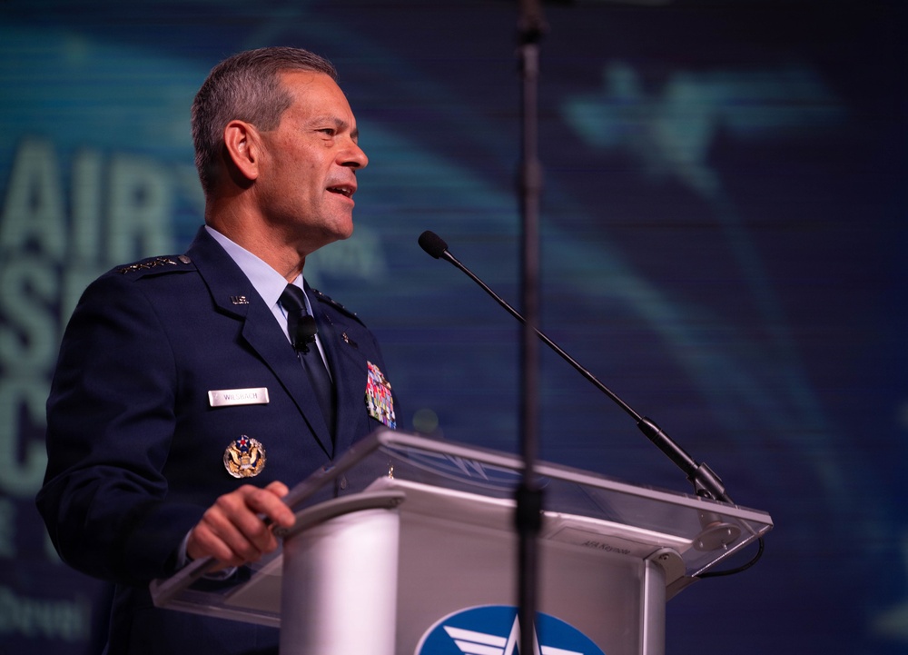 A senior U.S. Air Force officer in dress uniform is delivering a speech at a podium equipped with two microphones. The background displays partial text reading 'AIR' and what appears to be an aviation-themed conference backdrop with aircraft silhouettes. The setting appears to be a formal military or aerospace industry conference.