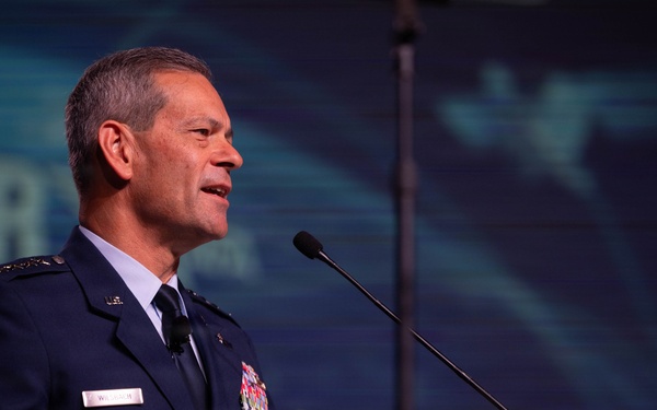 A senior U.S. Air Force officer in dress uniform is delivering a speech at a podium equipped with two microphones. The background displays partial text reading 'AIR' and what appears to be an aviation-themed conference backdrop with aircraft silhouettes. The setting appears to be a formal military or aerospace industry conference.