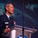 A senior U.S. Air Force officer in dress uniform is delivering a speech at a podium equipped with two microphones. The background displays partial text reading 'AIR' and what appears to be an aviation-themed conference backdrop with aircraft silhouettes. The setting appears to be a formal military or aerospace industry conference.