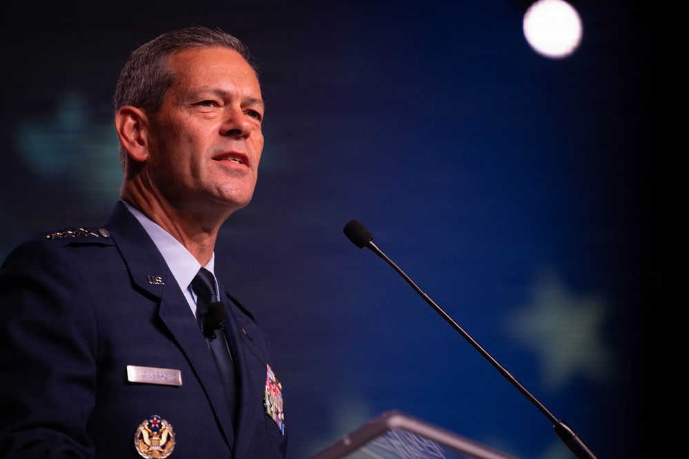 A senior U.S. Air Force officer in dress uniform is speaking at a podium with a microphone. He is wearing a dark blue service dress uniform with multiple medal ribbons, rank insignia on his shoulders, and a name tag. The background features a dark blue stage setting with subtle star-like design elements.