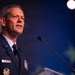 A senior U.S. Air Force officer in dress uniform is speaking at a podium with a microphone. He is wearing a dark blue service dress uniform with multiple medal ribbons, rank insignia on his shoulders, and a name tag. The background features a dark blue stage setting with subtle star-like design elements.