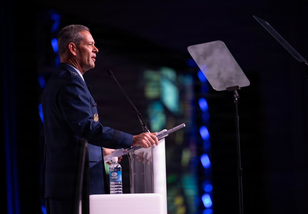 A military officer in a dark blue dress uniform is delivering a speech at a podium on a stage. The setting appears to be a formal conference or ceremony with professional stage lighting. A music stand or teleprompter stand is visible to the right of the speaker.