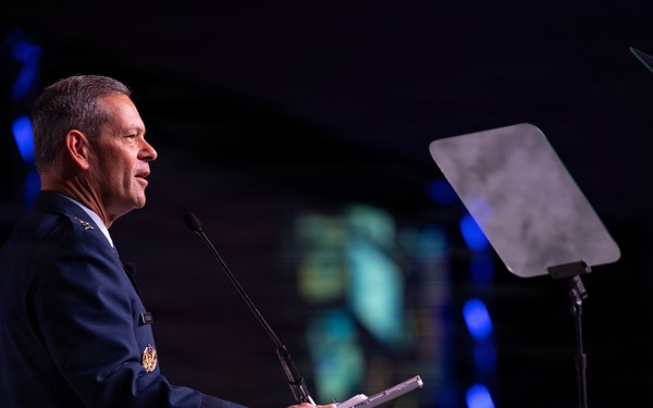 A military officer in a dark blue dress uniform is delivering a speech at a podium on a stage. The setting appears to be a formal conference or ceremony with professional stage lighting. A music stand or teleprompter stand is visible to the right of the speaker.