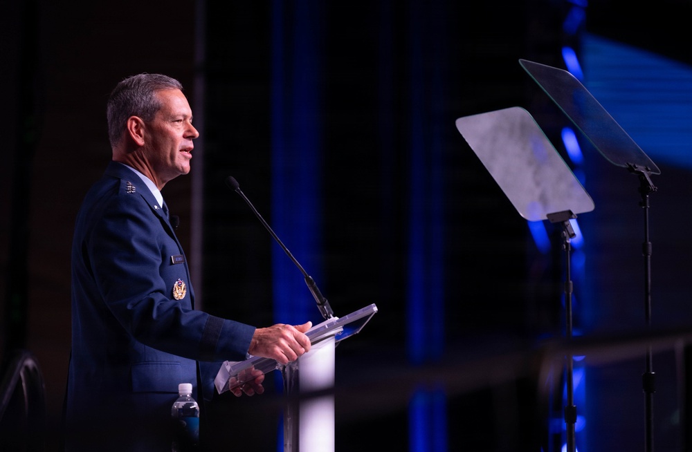 A senior military officer in a blue dress uniform is delivering a speech at a podium equipped with a microphone. The setting appears to be a formal conference or ceremonial event, indicated by the professional stage lighting and teleprompter visible to the right. The officer's uniform displays multiple insignia and medals consistent with a high-ranking position.
