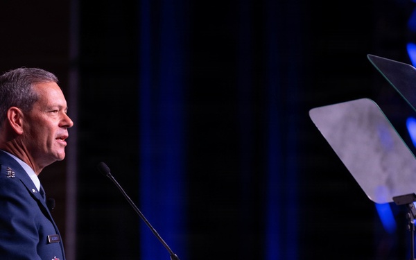 A senior military officer in a blue dress uniform is delivering a speech at a podium equipped with a microphone. The setting appears to be a formal conference or ceremonial event, indicated by the professional stage lighting and teleprompter visible to the right. The officer's uniform displays multiple insignia and medals consistent with a high-ranking position.