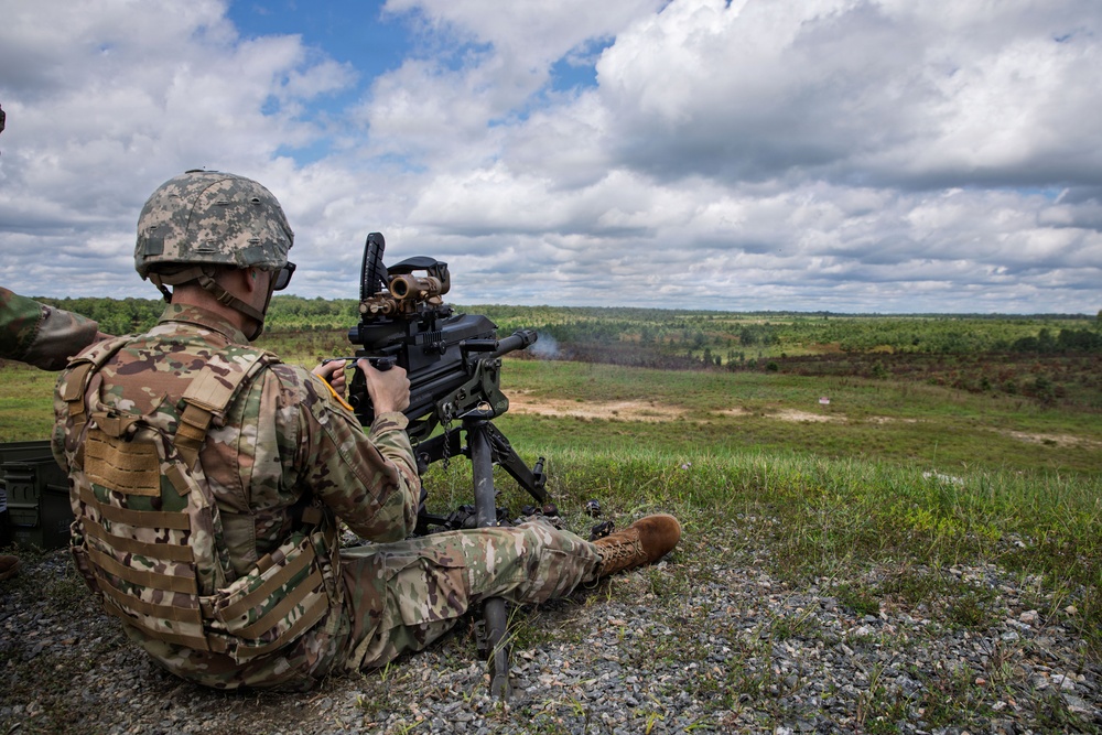 A soldier in U.S. Army MultiCam/OCP camouflage uniform is seated on a gravel berm operating a crew-served weapon, appearing to be an M19 or MK19 grenade launcher mounted on a tripod. Smoke is visible at the muzzle, indicating the weapon has just been fired. The setting appears to be a military training range with an open grassy field downrange under a partly cloudy sky.