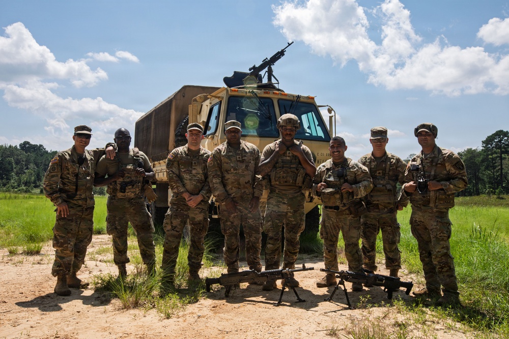 A group of approximately eight U.S. Army soldiers in MultiCam/OCP camouflage uniforms pose for a group photo in front of a tan military tactical truck (LMTV/FMTV) on a dirt field. Two crew-served weapons (likely M240 machine guns) are positioned on bipods in the foreground, and a mounted weapon is visible on top of the vehicle. The setting appears to be a training range or field exercise area with open grassland and tree line in the background.