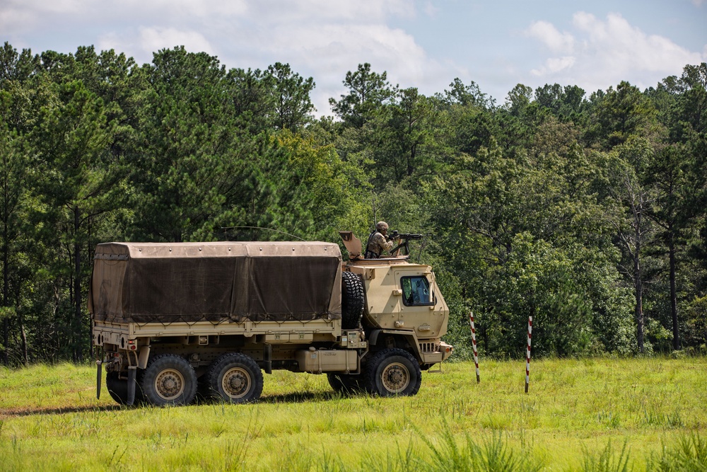 A tan/desert-camouflage military tactical truck, consistent with an FMTV (Family of Medium Tactical Vehicles), is positioned in an open grassy field at the edge of a tree line. A soldier is visible in a turret position atop the cab, appearing to man a weapon system. Red and white marker poles are visible to the right of the vehicle.
