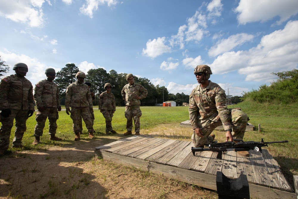 A group of military personnel in U.S. Army camouflage (OCP/MultiCam) uniforms are gathered at an outdoor firing range. One soldier, kneeling on a wooden platform, appears to be conducting a weapons demonstration or instruction with what appears to be an M249 SAW (Squad Automatic Weapon) or similar light machine gun. The remaining soldiers stand in a line observing the instruction.
