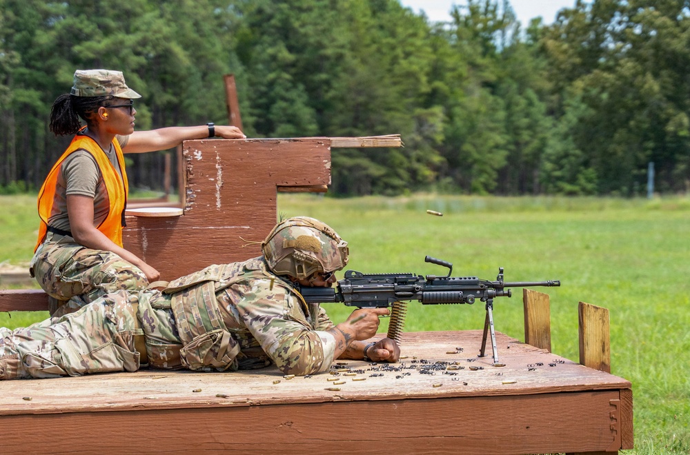 Two military personnel are engaged in a live-fire training exercise at an outdoor shooting range. One soldier in full combat gear and helmet is in the prone position operating what appears to be an M249 Squad Automatic Weapon (SAW) on a wooden platform, with spent brass casings visible. A second soldier wearing an orange safety vest is acting as a range safety officer or observer, monitoring the exercise.