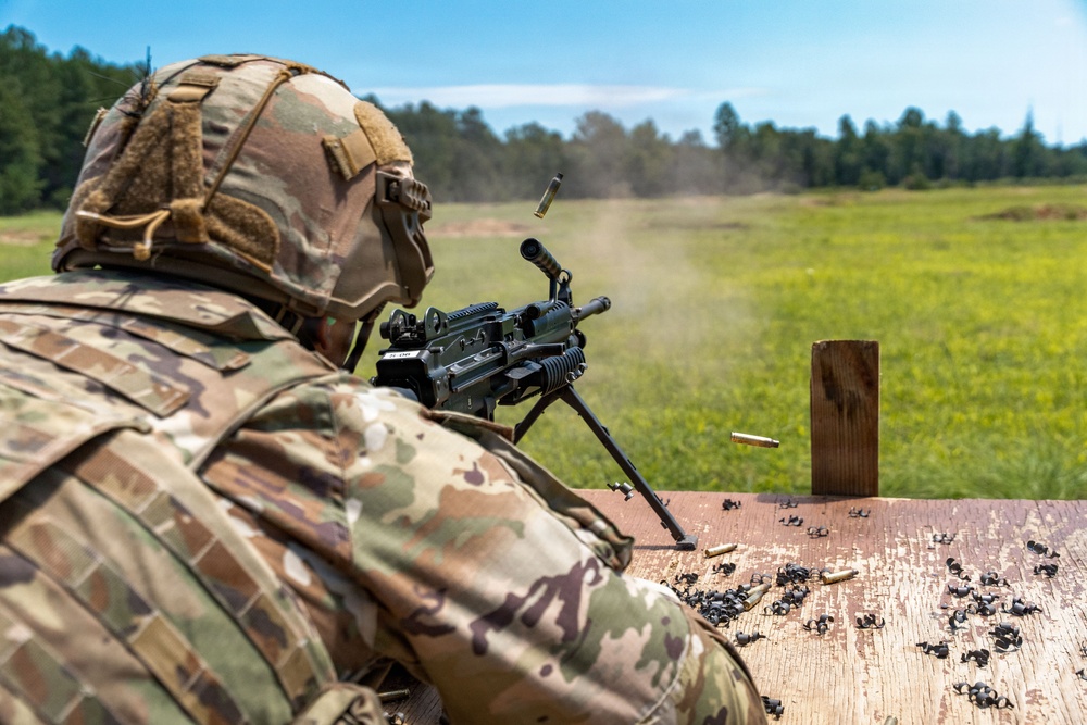 A soldier in full camouflage uniform and tactical helmet is firing a light machine gun mounted on a bipod from a wooden shooting bench at an outdoor range. Multiple spent brass casings are visible mid-air and scattered across the bench surface, indicating active firing. The setting appears to be a military training range with an open grass field and tree line in the background.