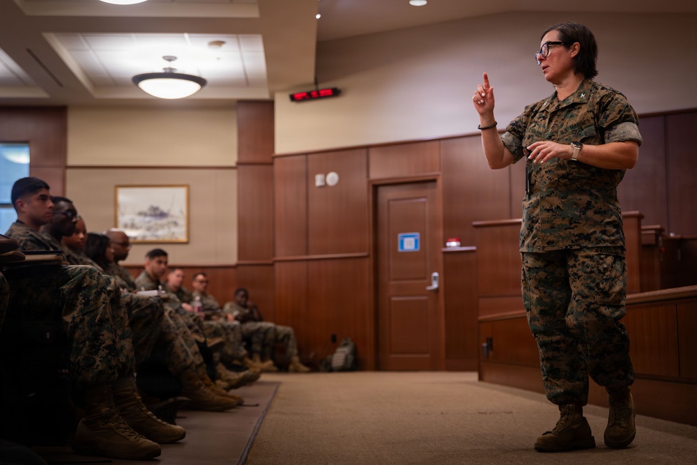 A female military officer in U.S. Marine Corps MARPAT camouflage uniform is addressing a group of seated Marines in what appears to be a formal briefing room or auditorium. She is standing and gesturing with one finger raised, suggesting she is making a key point during a presentation or instruction session. The audience of Marines is seated along the left side of the room, listening attentively.