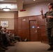 A female military officer in U.S. Marine Corps MARPAT camouflage uniform is addressing a group of seated Marines in what appears to be a formal briefing room or auditorium. She is standing and gesturing with one finger raised, suggesting she is making a key point during a presentation or instruction session. The audience of Marines is seated along the left side of the room, listening attentively.