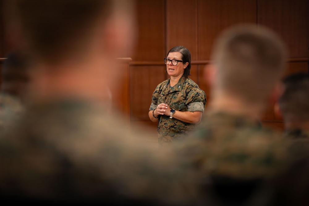 A female military officer in U.S. Marine Corps MARPAT camouflage uniform is addressing a group of seated Marines in what appears to be a formal briefing or meeting room. She is standing at the front of the room with her hands clasped, speaking to an audience whose backs and heads are visible in the foreground. The setting suggests an official military briefing, training session, or command presentation.