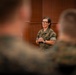 A female military officer in U.S. Marine Corps MARPAT camouflage uniform is addressing a group of seated Marines in what appears to be a formal briefing or meeting room. She is standing at the front of the room with her hands clasped, speaking to an audience whose backs and heads are visible in the foreground. The setting suggests an official military briefing, training session, or command presentation.