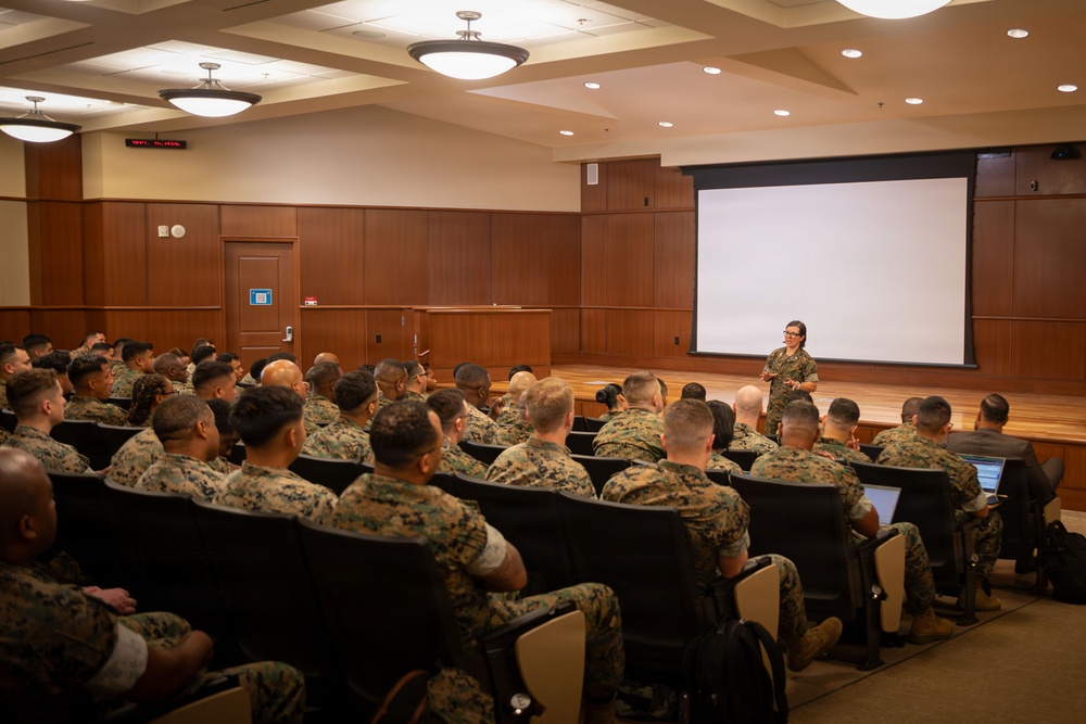 A female military instructor or officer in camouflage uniform is presenting to a large audience of uniformed military personnel in a formal briefing room. The audience, seated in rows of chairs, appears to be U.S. Marines based on their distinctive MARPAT (Marine Pattern) camouflage uniforms. A large projection screen is visible at the front of the room, though no content is currently displayed on it.