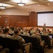 A female military instructor or officer in camouflage uniform is presenting to a large audience of uniformed military personnel in a formal briefing room. The audience, seated in rows of chairs, appears to be U.S. Marines based on their distinctive MARPAT (Marine Pattern) camouflage uniforms. A large projection screen is visible at the front of the room, though no content is currently displayed on it.