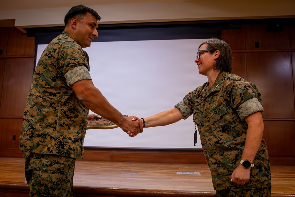 Two U.S. Marines in MARPAT woodland camouflage uniforms are shaking hands in what appears to be a formal ceremony or recognition event inside a military briefing room or auditorium. The individual on the left is holding what appears to be a plaque or award. The female Marine on the right displays a Lieutenant Colonel oak leaf cluster insignia on her collar.