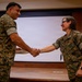 Two U.S. Marines in MARPAT woodland camouflage uniforms are shaking hands in what appears to be a formal ceremony or recognition event inside a military briefing room or auditorium. The individual on the left is holding what appears to be a plaque or award. The female Marine on the right displays a Lieutenant Colonel oak leaf cluster insignia on her collar.