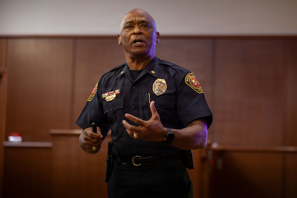 A law enforcement officer in a dark navy blue uniform is speaking or presenting in what appears to be a formal indoor setting such as a courtroom or conference room. He is gesturing with his right hand while holding an object in his left hand, suggesting he is actively addressing an audience. His uniform includes a badge, shoulder patches, rank insignia, and a name tag.