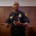 A law enforcement officer in a dark navy blue uniform is speaking or presenting in what appears to be a formal indoor setting such as a courtroom or conference room. He is gesturing with his right hand while holding an object in his left hand, suggesting he is actively addressing an audience. His uniform includes a badge, shoulder patches, rank insignia, and a name tag.