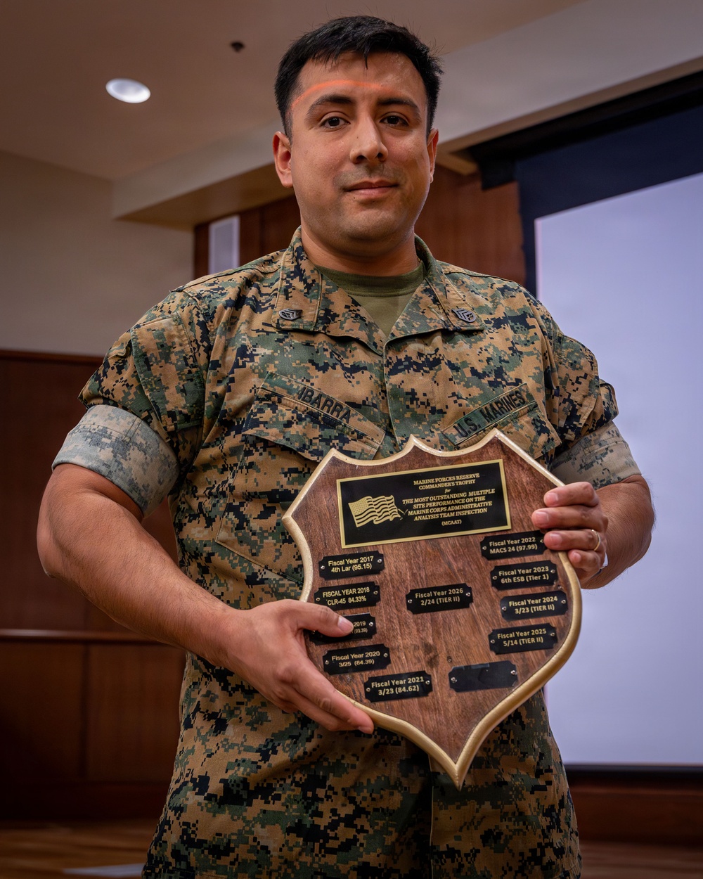 A U.S. Marine in MARPAT digital camouflage uniform is holding a wooden shield-shaped award plaque. The plaque is identified as the Marine Forces Reserve Commander's Trophy for the Most Outstanding Multiple Site Performance on the Marine Corps Administrative Analysis Team Inspection (MCAAT). The Marine's name tape reads 'IBARRA' and the branch tape reads 'U.S. MARINES'.