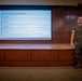 A military service member in MARPAT (Marine Corps digital camouflage) uniform stands in front of a projected presentation slide titled 'Four Process-Based Branches' in what appears to be a briefing room or auditorium. The slide outlines four organizational branches: Maintenance, Separations, Travel &amp; Ops, and Systems &amp; QA. The setting suggests a formal military administrative briefing or training session.