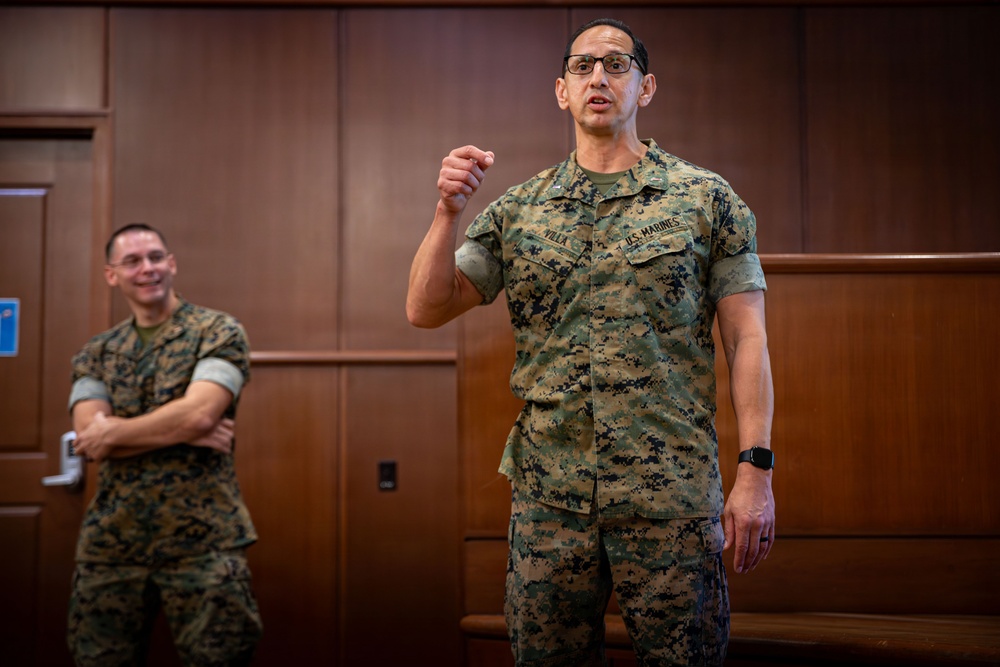 Two U.S. Marines in MARPAT (Marine Pattern) camouflage uniforms are shown in what appears to be a briefing or meeting room with wood-paneled walls. The Marine in the foreground is speaking and gesturing with a raised fist, while the Marine in the background stands with arms crossed and is smiling. The name tape on the foreground Marine reads 'VILLA' and the branch tape reads 'U.S. MARINES'.