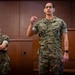 Two U.S. Marines in MARPAT (Marine Pattern) camouflage uniforms are shown in what appears to be a briefing or meeting room with wood-paneled walls. The Marine in the foreground is speaking and gesturing with a raised fist, while the Marine in the background stands with arms crossed and is smiling. The name tape on the foreground Marine reads 'VILLA' and the branch tape reads 'U.S. MARINES'.