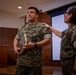 Two U.S. Marines in MARPAT woodland camouflage uniforms are participating in what appears to be an award or recognition ceremony indoors. The Marine on the left is holding a wooden plaque or award, while the Marine on the right has a hand placed on their shoulder in a congratulatory gesture. The setting appears to be a formal meeting or conference room with wood paneling and a projection screen in the background.