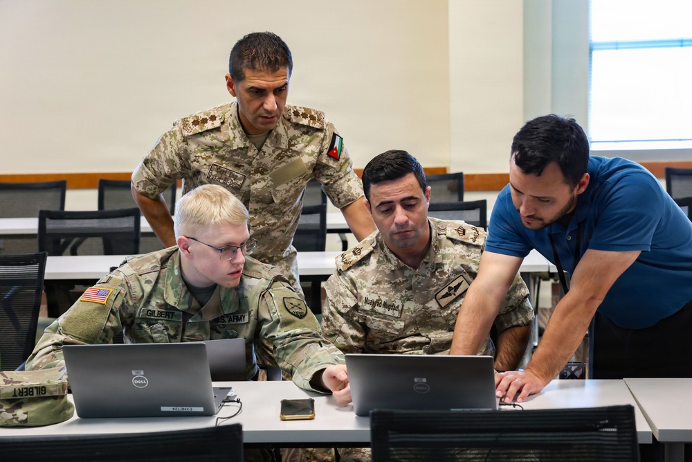 Four individuals are gathered around two Dell laptops in what appears to be a military training or briefing room. The group includes a U.S. Army soldier identified by name tape as 'Gilbert,' two personnel in desert camouflage uniforms bearing a flag patch consistent with Jordan, and one civilian in a blue polo shirt. They appear to be collaborating on a task displayed on the laptop screens.