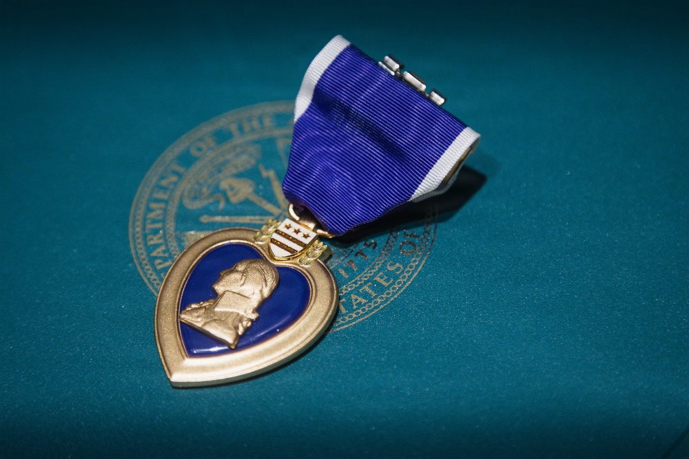 The image shows a United States Purple Heart military medal resting on a teal surface bearing a partial Department of Defense or military department seal. The medal features its characteristic heart-shaped design with a gold border, blue enamel center, and a profile relief of George Washington. It is attached to a purple and white striped ribbon with a suspension bar.
