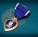 The image shows a United States Purple Heart military medal resting on a teal surface bearing a partial Department of Defense or military department seal. The medal features its characteristic heart-shaped design with a gold border, blue enamel center, and a profile relief of George Washington. It is attached to a purple and white striped ribbon with a suspension bar.