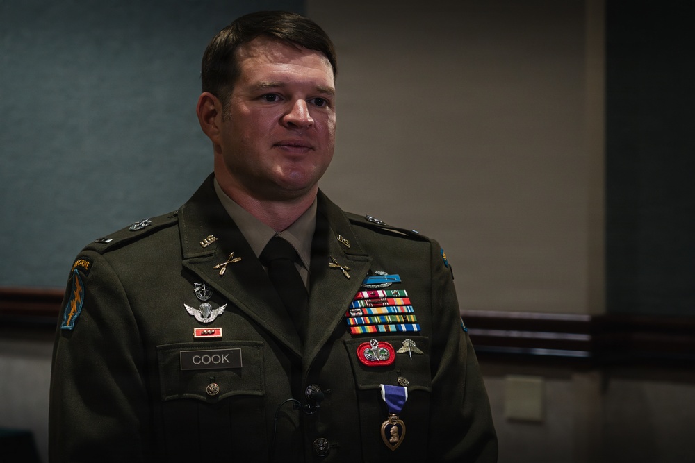 A U.S. Army soldier named Cook, wearing Army Service Uniform (ASU) in olive green, is standing at attention or in a formal posture during what appears to be a military ceremony. He displays numerous decorations including a Purple Heart medal, multiple service ribbons, airborne wings, and special forces insignia. The 'AIRBORNE' tab and unit patches are visible on his left shoulder.