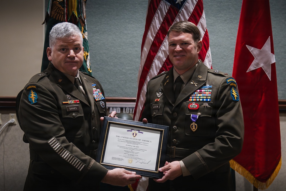 Two U.S. Army soldiers in dress green uniforms are posing together holding a framed Purple Heart certificate during a formal military award ceremony. The soldier on the right is wearing a Purple Heart medal on his chest, indicating he is the recipient. Both soldiers display Special Forces (Airborne) insignia and extensive service ribbons.