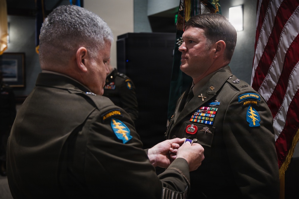 A military award ceremony is taking place in which one U.S. Army officer is pinning a medal onto another officer's uniform. The recipient is wearing a Special Forces Airborne uniform with numerous ribbons, badges, and unit patches. An American flag is visible in the background.